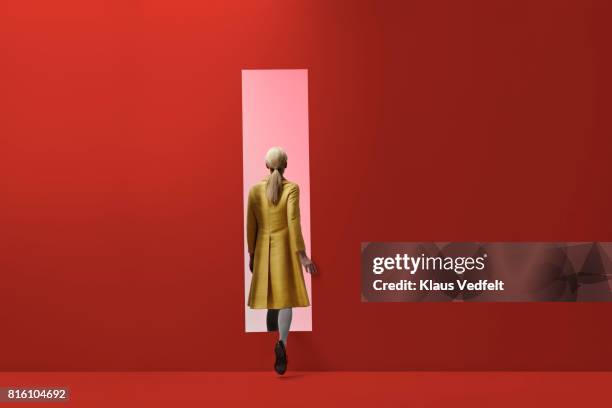 woman walking into rectangular opening in coloured wall - gele overjas stockfoto's en -beelden