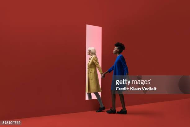 two women holding hands, walking threw rectangular opening in coloured wall - gele overjas stockfoto's en -beelden