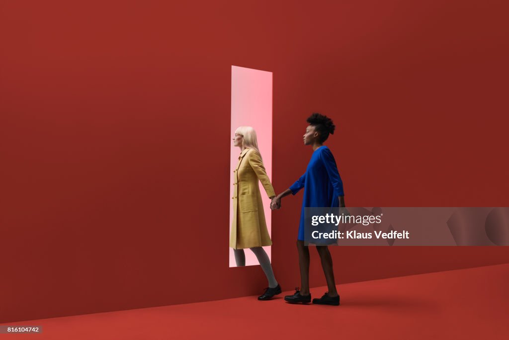 Two women holding hands, walking threw rectangular opening in coloured wall