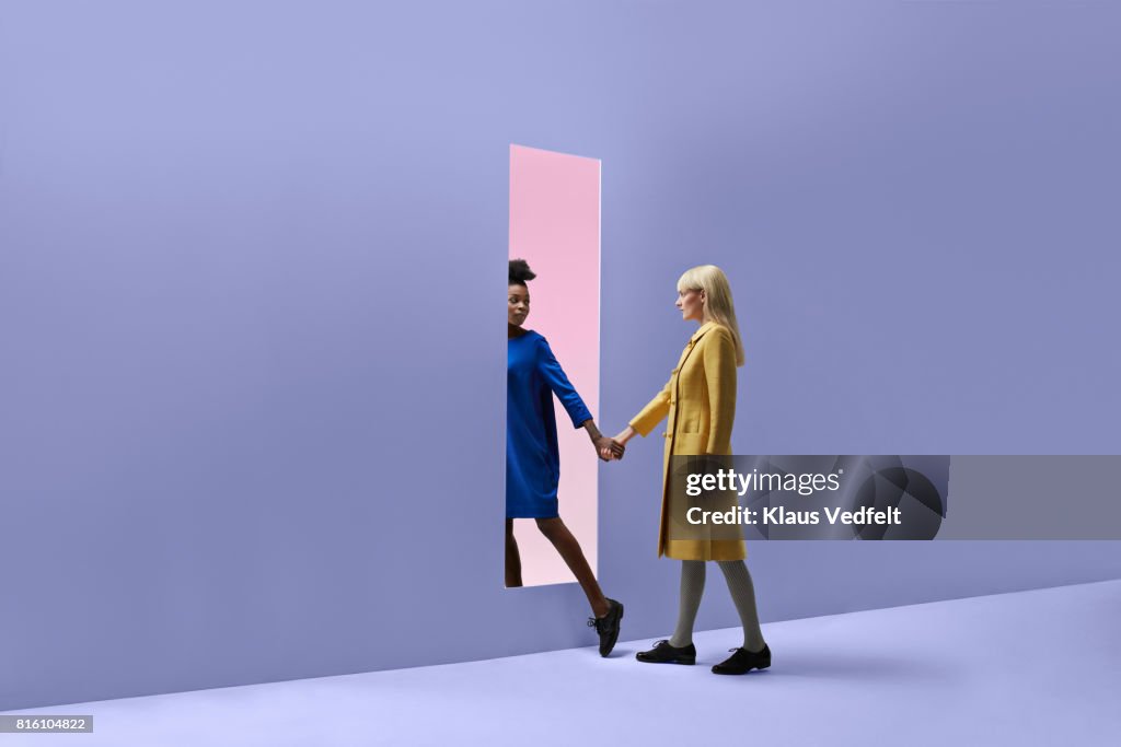Two women holding hands, walking threw rectangular opening in coloured wall