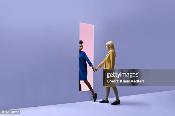two women holding hands, walking threw rectangular opening in coloured wall - viola colore foto e immagini stock