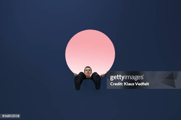 man sitting on edge of round opening in coloured ceiling, looking down with feet dangling - männerrunde stock-fotos und bilder