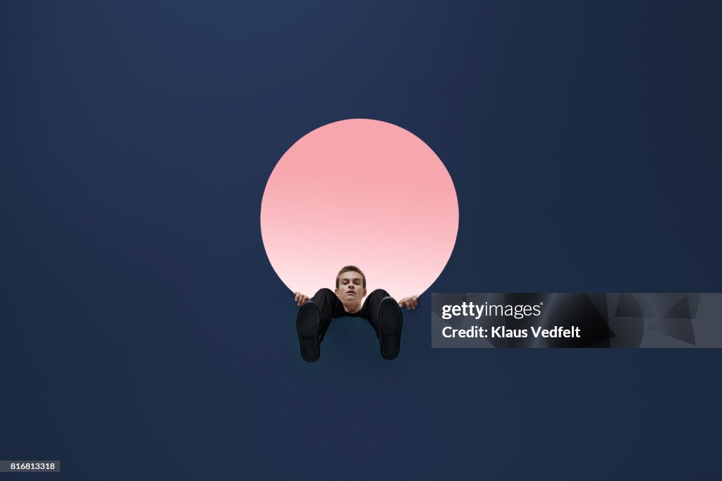 Man sitting on edge of round opening in coloured ceiling, looking down with feet dangling