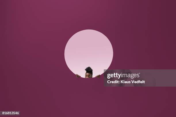 woman peeking out of round opening in coloured wall - unveiling foto e immagini stock