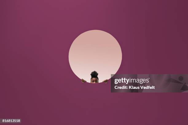 woman peeking out of round opening in coloured wall - spähen stock-fotos und bilder