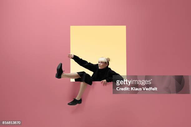 woman hanging on to square opening in coloured wall, feet dangling - appearance foto e immagini stock