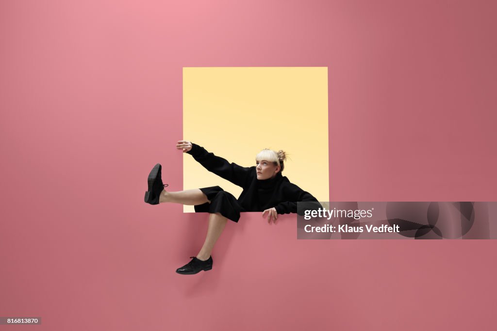 Woman hanging on to square opening in coloured wall, feet dangling