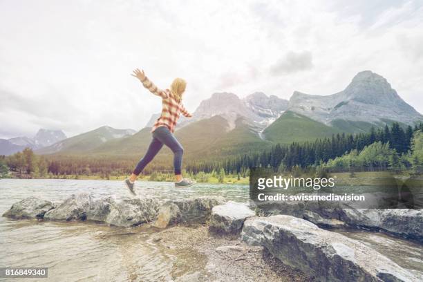 young woman jumps rock to rock on mountain lake - leap forward stock pictures, royalty-free photos & images