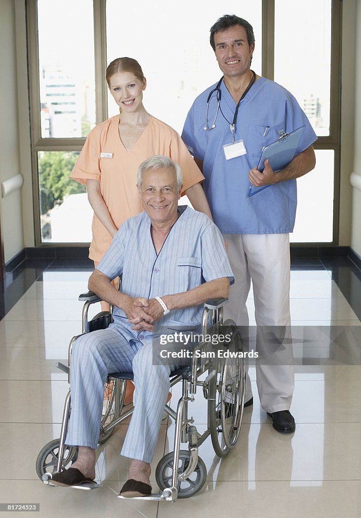 Two hospital workers in corridor with senior patient in wheelchair smiling