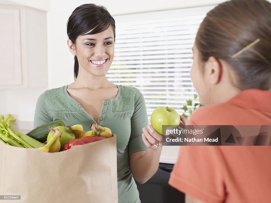 Woman handing young girl in kitchen an apple out of grocery bag and smiling