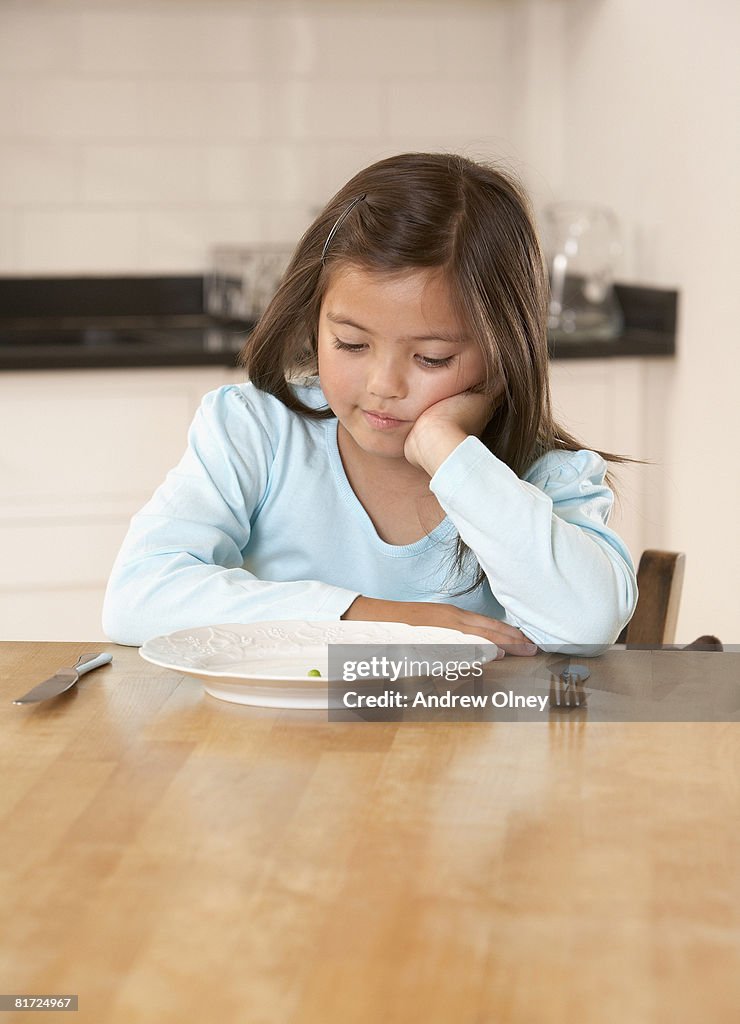 Young girl in kitchen with a single green pea on her plate looking unhappy