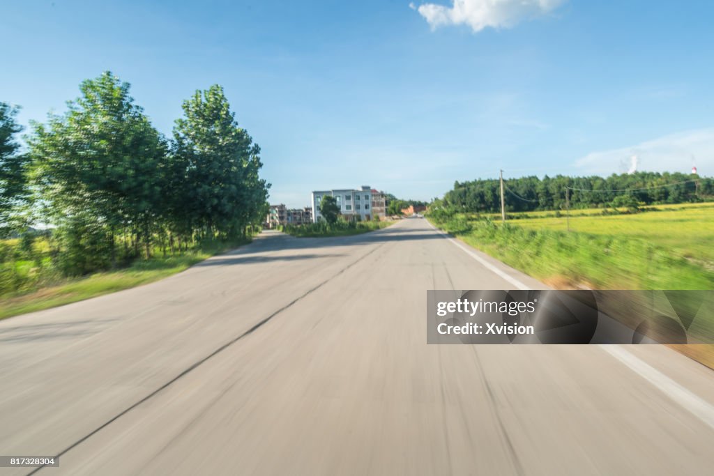 Asphalt road under blue sky with clouds in motion blur with plants in sides