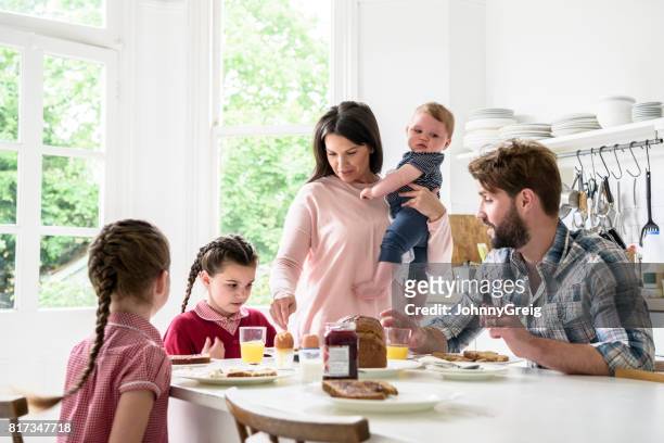 familie sitzt am frühstückstisch, mutter mit baby, vater und mädchen essen - busy mom stock-fotos und bilder
