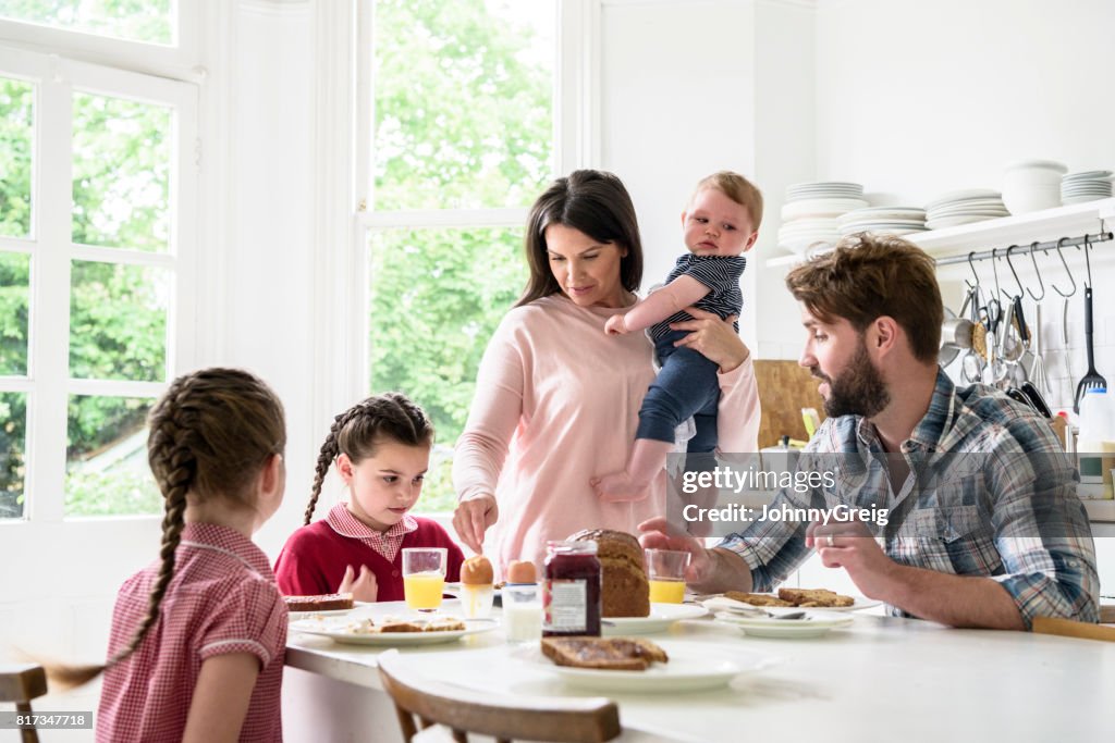 Family sitting at breakfast table, mother holding baby boy, father and girls eating food