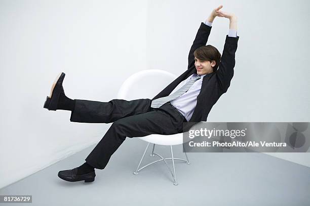 young man in suit sitting in chair, legs outstretched, arms raised, full length - mann sitzt auf stuhl stock-fotos und bilder