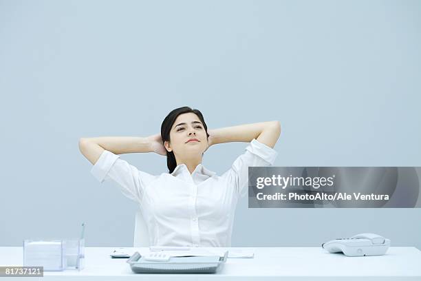 young woman sitting at desk with hands behind head, looking away, smiling - grijze bloes stockfoto's en -beelden