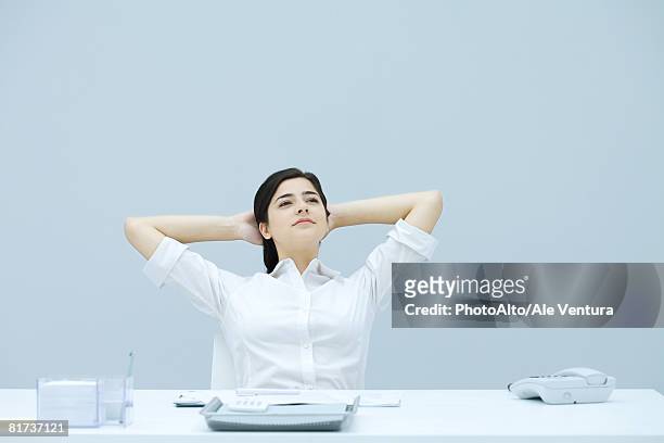 young woman sitting at desk with hands behind head, looking away, smiling - hands behind head stock pictures, royalty-free photos & images