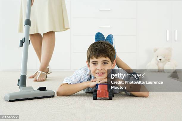 little boy lying on the ground with toys, smiling at camera, mother vacuuming around him - allongé sur le devant photos et images de collection