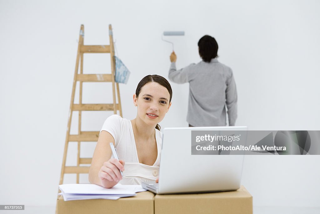 Young woman sitting at makeshift desk, using laptop computer, smiling at camera, man painting wall in background