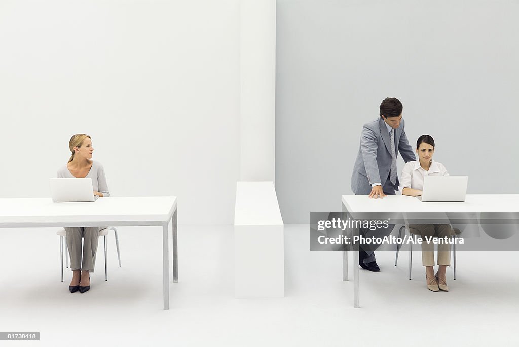 Two women working in office, man standing behind one with his hand on her shoulder