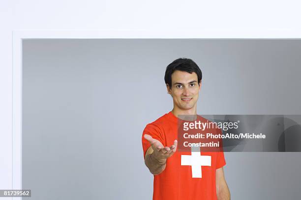young man wearing tee-shirt with plus symbol, holding out hand, smiling at camera - croce rossa foto e immagini stock