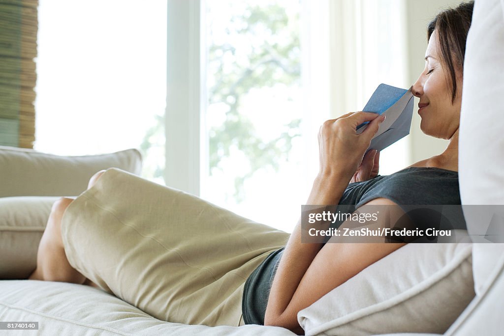 Woman sitting on sofa, holding up card and envelope to nose, smiling with eyes closed