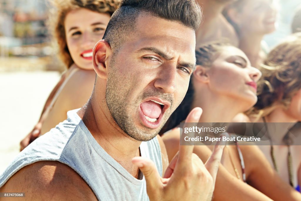 Young man with facial fair at the beach with friends on a sunny day making hand sign with mouth open