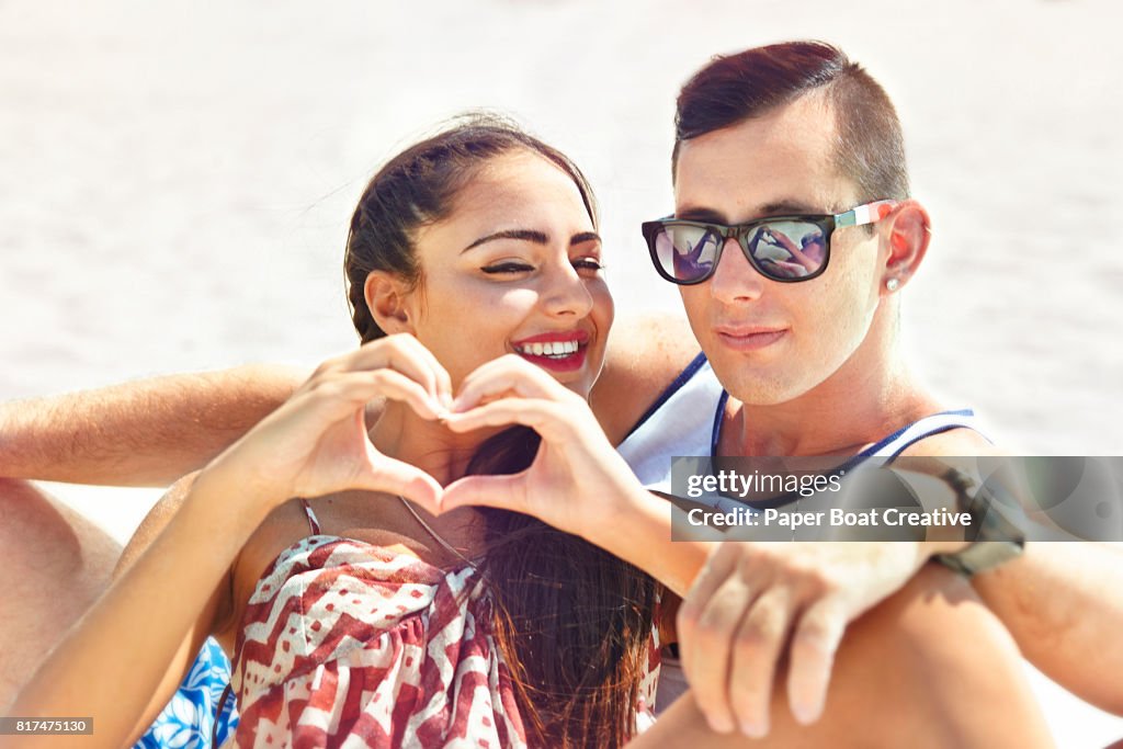 Handsome young couple sitting in the sand at the beach on a sunny day with heart sign