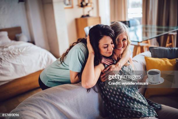 beautiful adult daughter embracing smiling senior mother sitting on sofa - mother daughter couch imagens e fotografias de stock