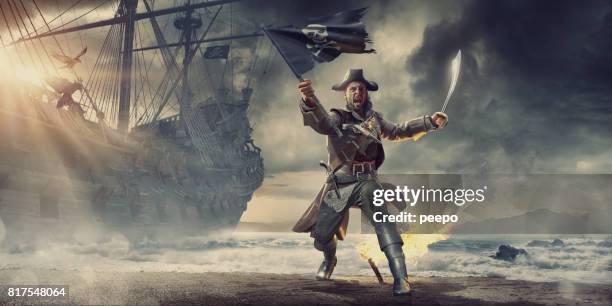 pirate on beach holding flag and cutlass near pirate ship - pirate stock pictures, royalty-free photos & images