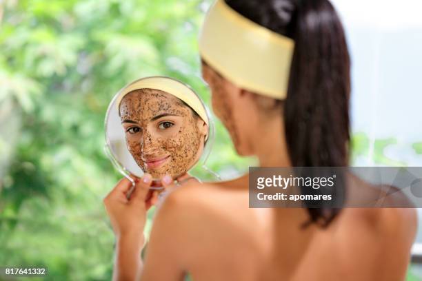hispanic young woman with coffe face scrub after bath looking at herself on hand mirror. skin exfoliation. - body scrub stock pictures, royalty-free photos & images