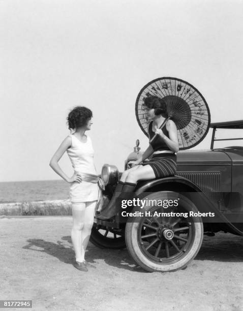 pair of women in old fashioned bathing suits at beach one sitting on car with oriental umbrella other standing in front of car talking to her. - 1920 stock-fotos und bilder