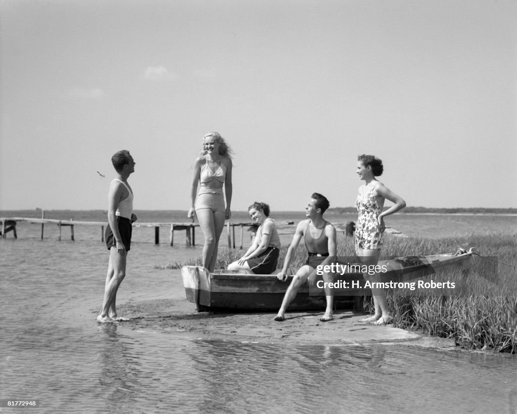 2 Men & 3 Women In Bathing Suits Grouped Around Beached Row Boat On Salt Marsh Smiling Dock In Background With Skyline Seashore Leisure Sandbar Summer.