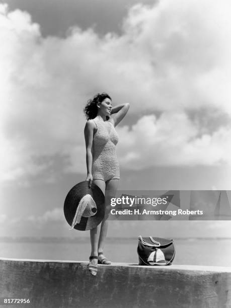 woman wearing knit bathing suit standing wall ocean sea background holding sun hat beach bag sail boat fashion beauty style swim wear retro. - fashion swimwear stock pictures, royalty-free photos & images