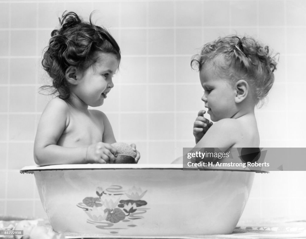 Two Baby Girls In One Tub One Is Holding A Sponge The Other Has Hand Raised To Mouth Both Are Naked Inside In The 1960s.