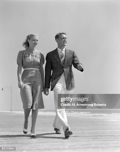 smiling couple walking on the beach she is wearing a shirt front dress with a white belt & spectator pumps he's wearing a shirt tie sport jacket & pants. - 1940 個照片及圖片檔