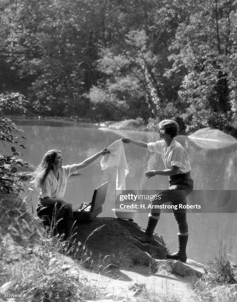 Two Women Camping By Lake Washing Clothes One Sitting On Rock The Other Standing Each Holding Peice Of Clothing Between Them Over Suitcase And Bowl.