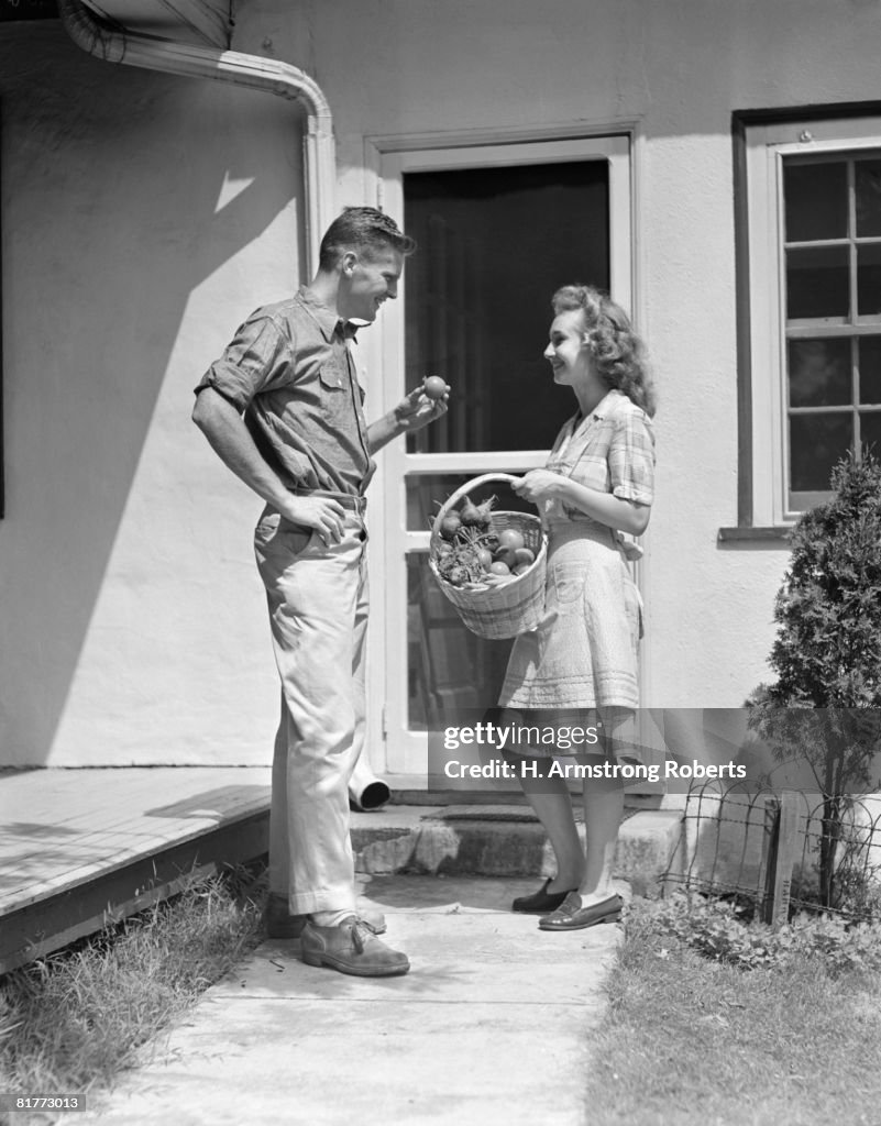 Smiling Couple Wearing A Workshirt & Chinos A Plaid Cotton Dress & Apron She Is Holding A Basket Of Vegetables He Is Holding A Tomato Standing At Doorway.