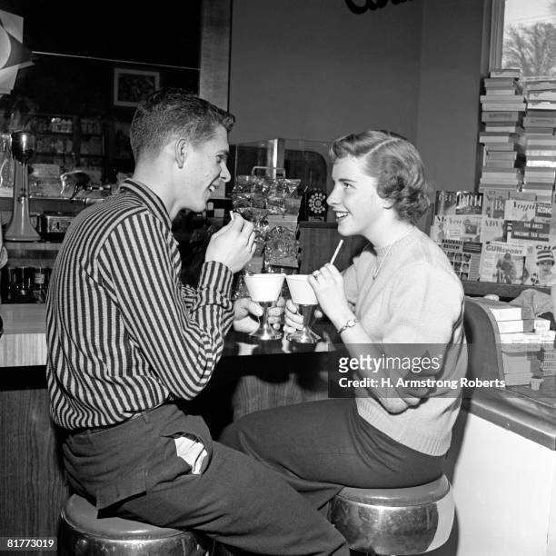 teen couple on stools at soda fountain drinking shakes & smiling at each other. - tomando sorvete imagens e fotografias de stock