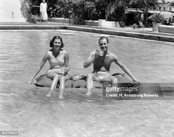 couple sitting on a float in a swimming pool wearing bathing suits holding drinks in thier hands smiling spectator serving tray sparkle. - women swimming pool retro stock pictures, royalty-free photos & images