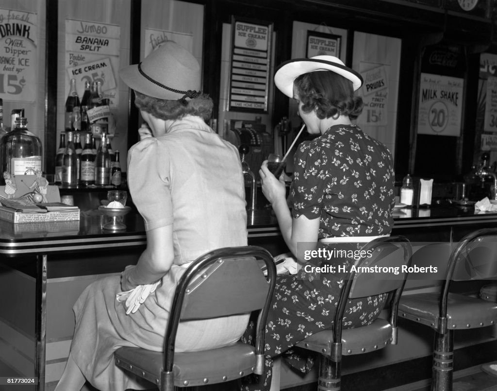 Back View Of 2 Women Both Wearing Hats 1 Wearing A Black & White Print Dress Eating Icecream & Sipping A Soda While Sitting At The Lunch Counter Stool.