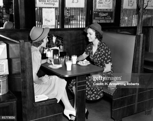 two smiling women both wearing hats 1 wearing dark print dress sitting in a booth having sandwiches & a glass of milk pumps signs water condiments. - 1940 個照片及圖片檔