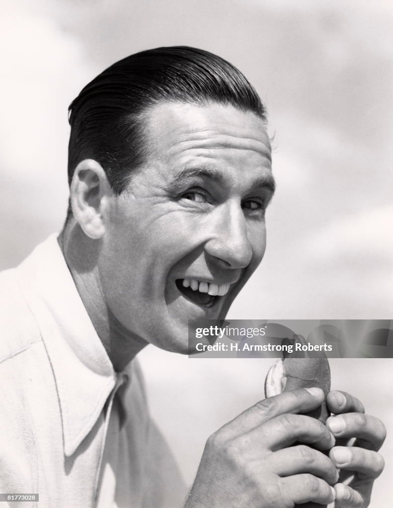 Head Shot Of Smiling Man With Wide Open Mouth And Head Pushed Forward About To Eat A Hotdog In A Frankfurter Roll Wearing A White Shirt Buck Teeth Hair.