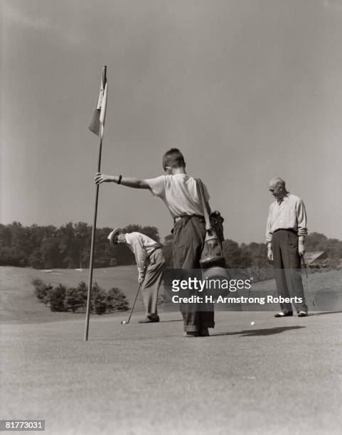 golfer getting ready to putt with boy caddy holding flag & partner looking on from side. - golf caddy stock-fotos und bilder