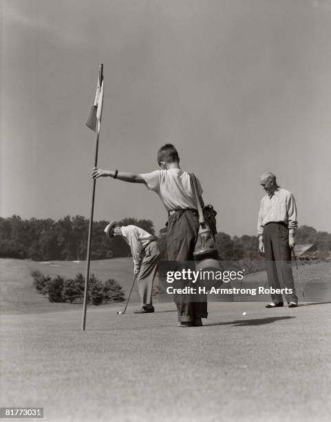 golfer getting ready to putt with boy caddy holding flag & partner looking on from side. - 20th century stock pictures, royalty-free photos & images