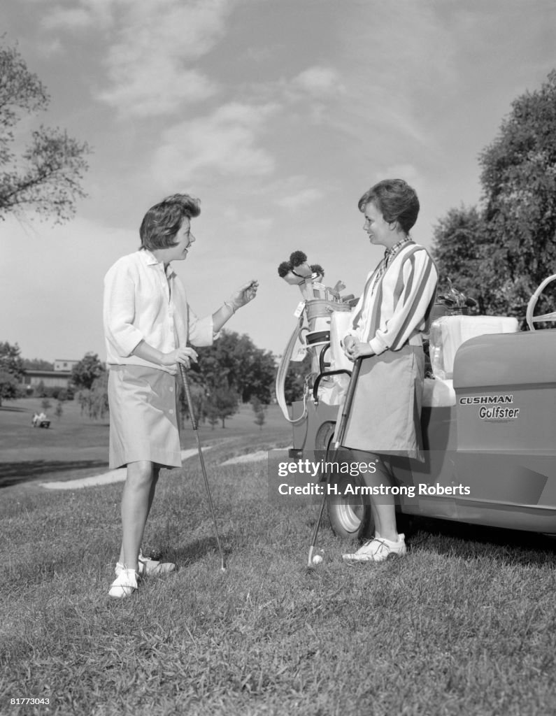 Pair Of Women On Golf Course Chatting One Leaning Against Golf Cart.