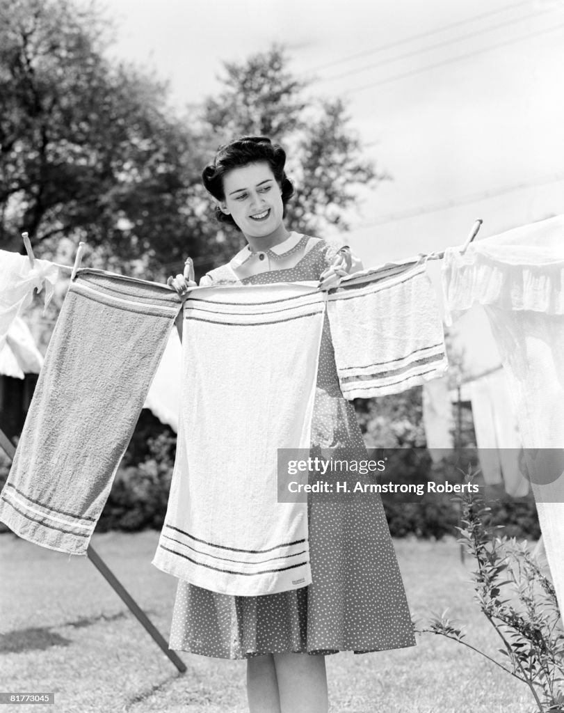 Woman Wearing A White Collar Poka Dot Cotton Dress Smiling While Pinning A Towel On A Clothes Line Clothes Prop Clothes Pin Foliage.