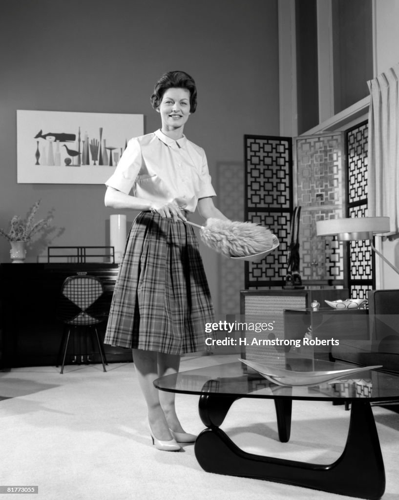 Woman Wearing A White Blouse & Plaid Skirt Dusting A Bowl With A Feather Duster In Front Of A Glass Top Coffee Table Smiling Modern Living Room.