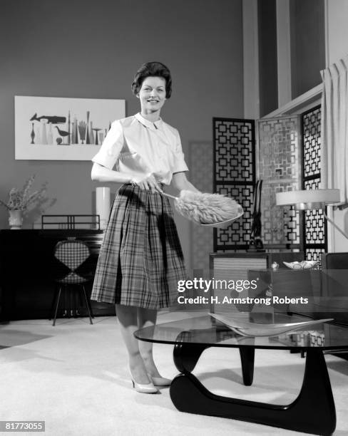 woman wearing a white blouse & plaid skirt dusting a bowl with a feather duster in front of a glass top coffee table smiling modern living room. - 1950s housewife stockfoto's en -beelden