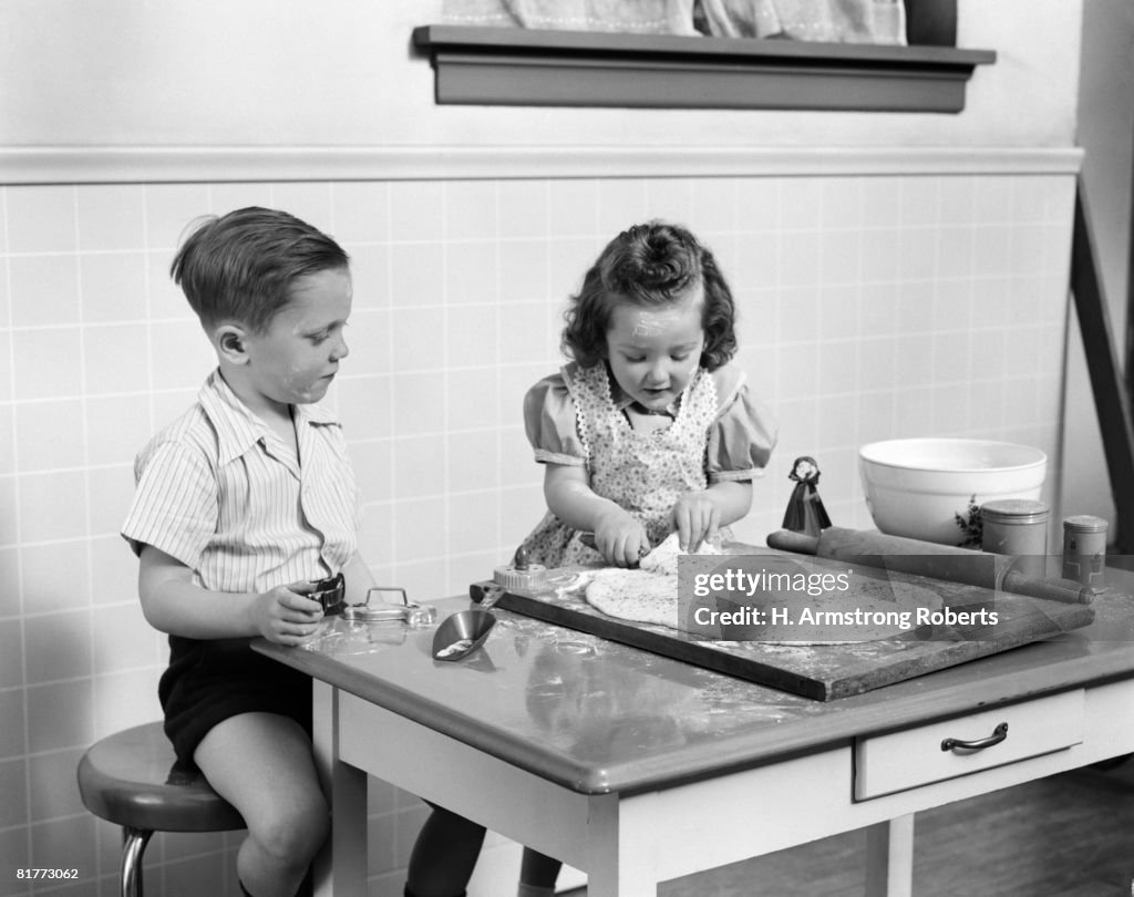 Boy In Shirt & Shorts Sitting At Table Watching Standing Girl In Print Apron Over Dress Preparing Cookie Dough Flour Rolling Pin Molds Baking Kitchen.